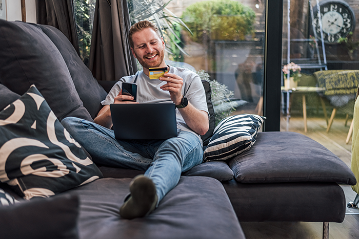 Man sitting on couch with laptop on his lap, holding his phone and a bank card and smiling
